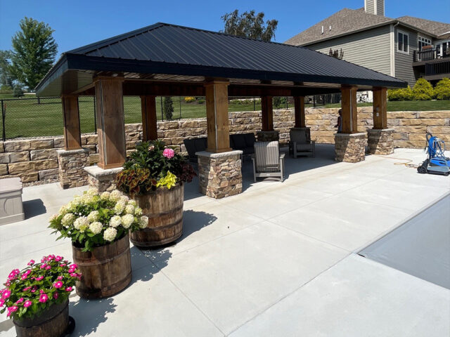 stone and wood pillar gazebo with metal roof and large retaining wall surrounging a pool