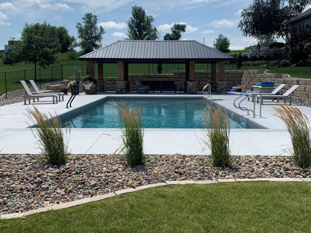 pool surrounded by concrete area bordered by river rock, plants and large retaining wall by gazebo